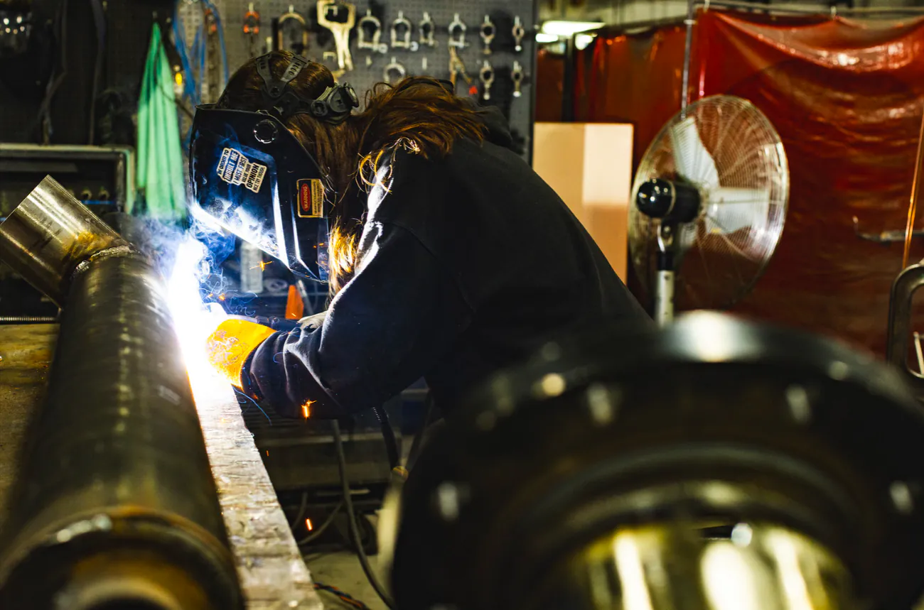 A welder in a protective helmet welds a steel pipe in a fabrication shop
